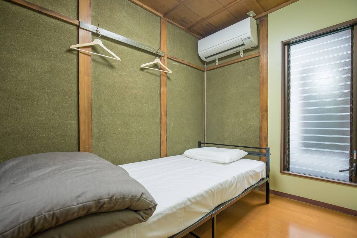 A single bed with a wooden frame, a white mattress, and a gray pillow. There is a wooden shelf with two white hangers and an air conditioning unit mounted on the wall. A window with blinds is visible to the right.