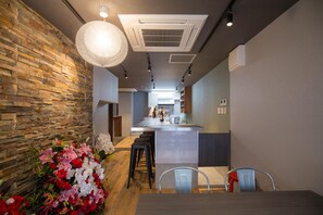 A modern kitchen with a central island, stone wall, and a floral arrangement. - Woodon - Hostel (Osaka)