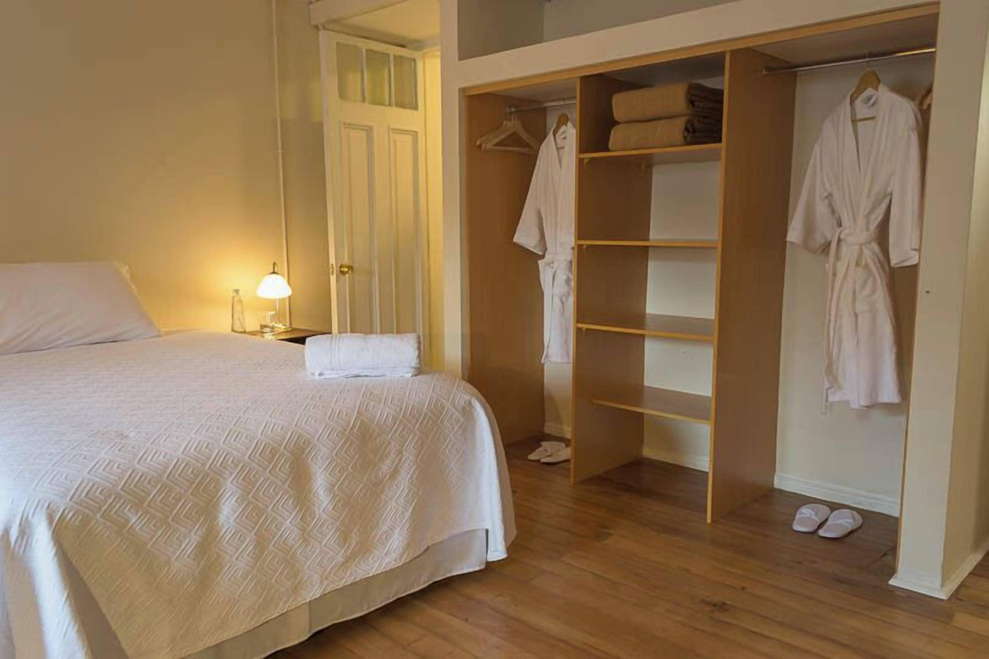 Ein Schlafzimmer mit Bett, Nachttischlampe und begehbarem Kleiderschrank mit Regalen und aufgehängten Bademänteln.
