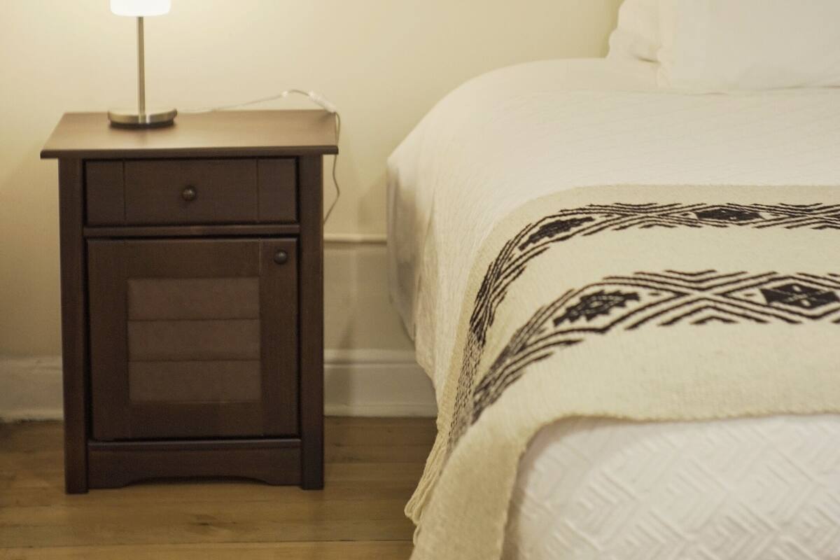 Un dormitorio con una mesita de noche de madera, una lámpara de noche, una cama con manta estampada y una pared lisa.
