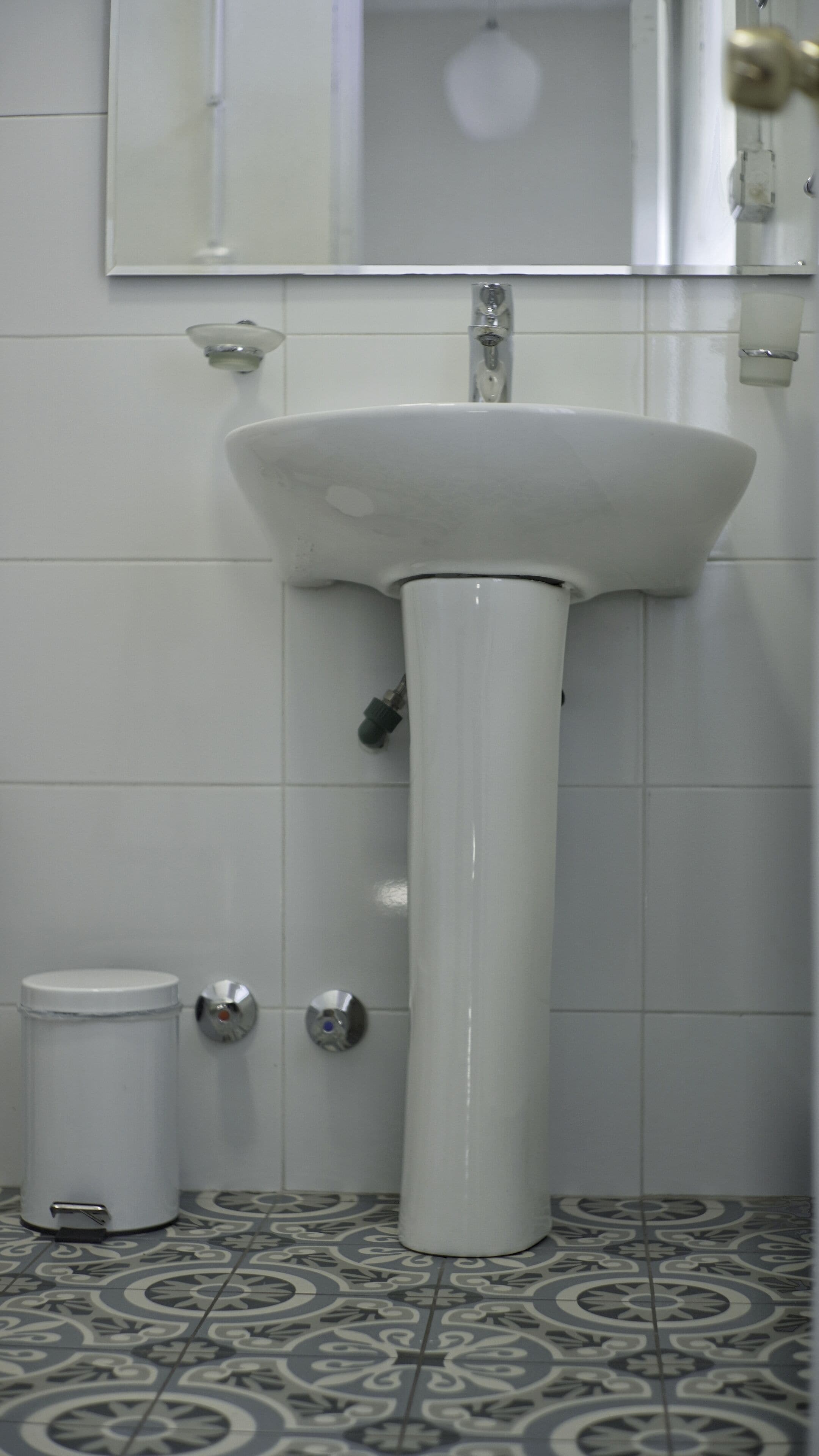 A bathroom with a white pedestal sink, a toilet, and a trash can. The floor has patterned tiles.