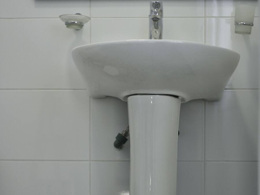 A bathroom with a white pedestal sink, a toilet, and a trash can. The floor has patterned tiles.