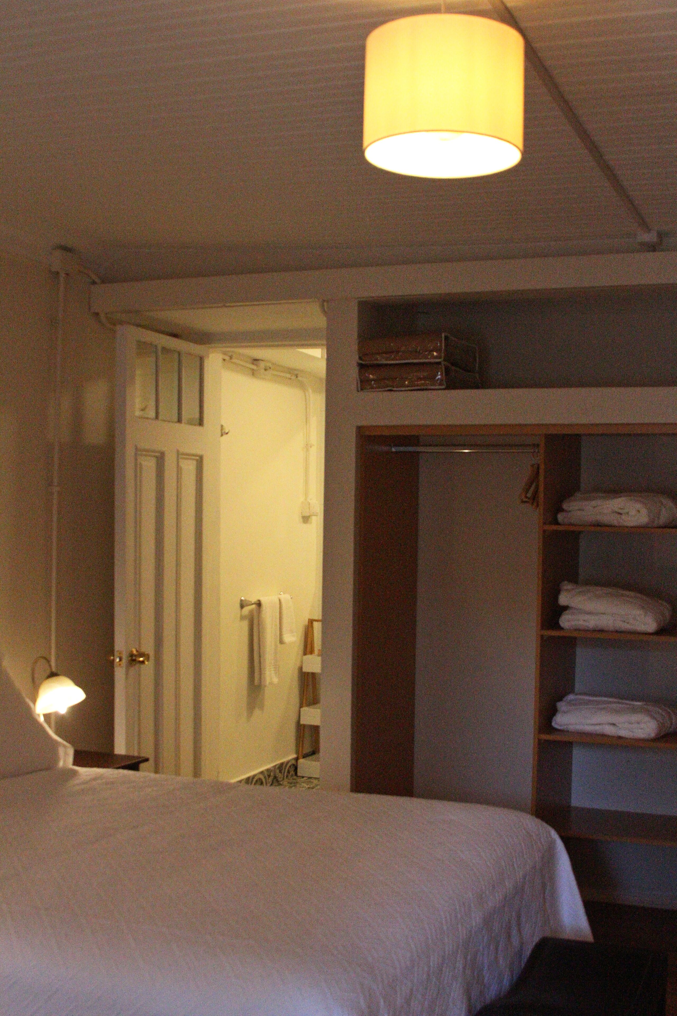 A hotel room with a bed, a bedside lamp, a wardrobe with shelves, and a door leading to a bathroom.