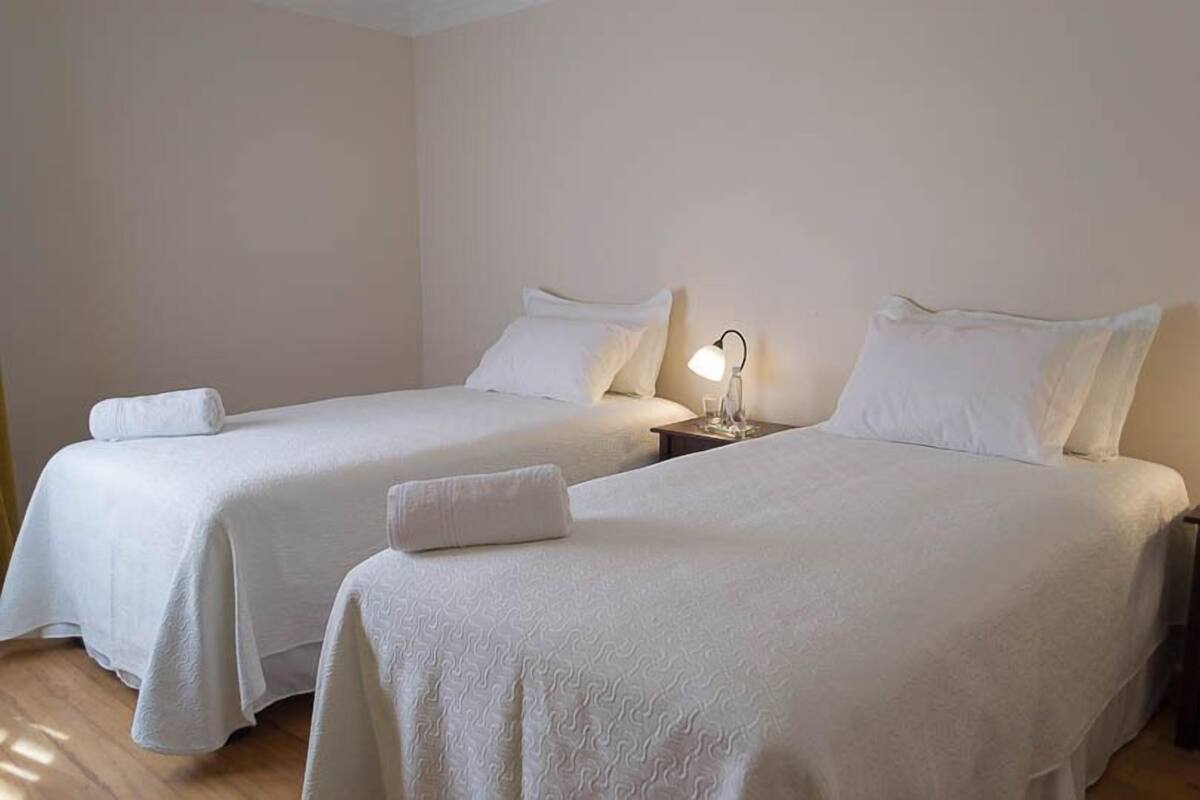 Dos camas individuales con sábanas y almohadas blancas, una mesita de noche con lámpara y suelo de madera.