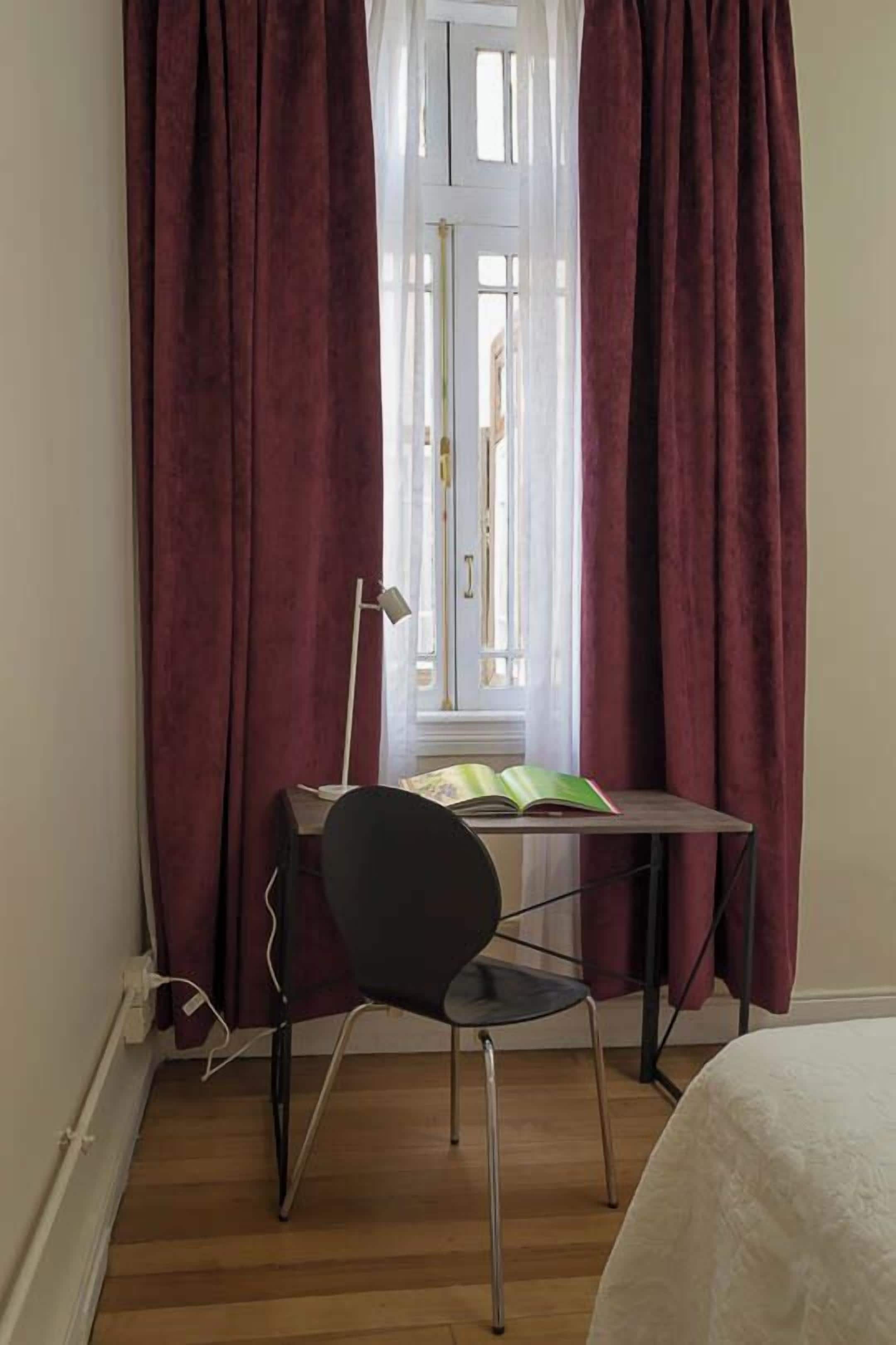 A room with a wooden floor, a desk with a chair, a lamp, and a bed.
