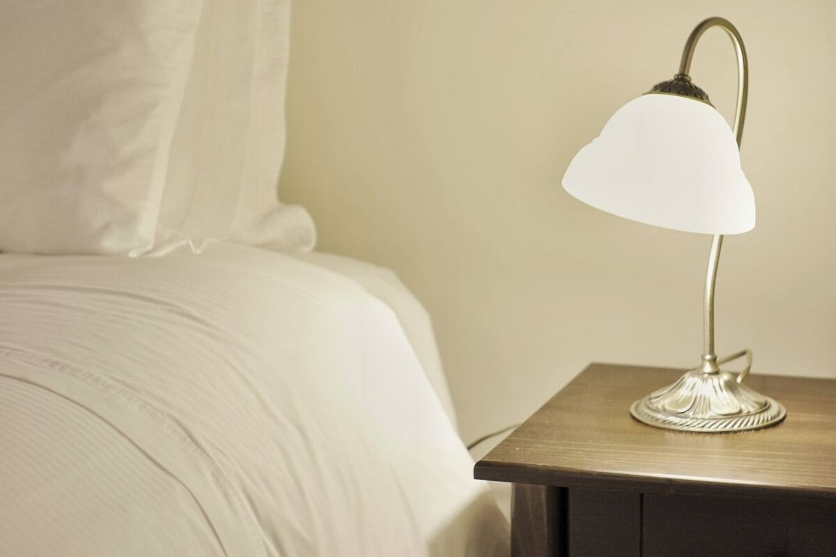 Una cama bien hecha con edredón blanco y almohadas, una mesita de noche con lámpara y una mesita auxiliar de madera oscura.