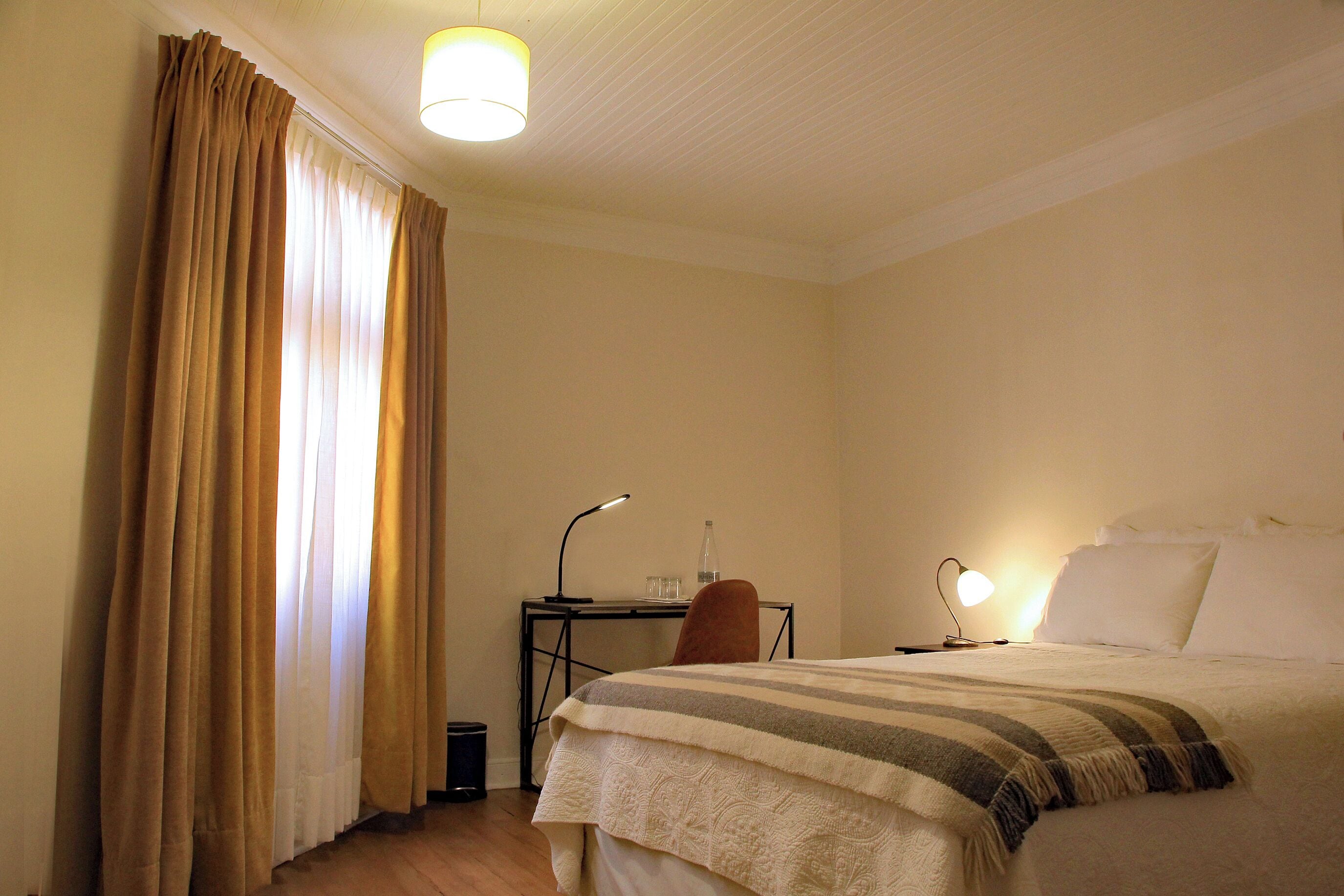A hotel room with a bed, a desk with a lamp, a chair, and a window with curtains.