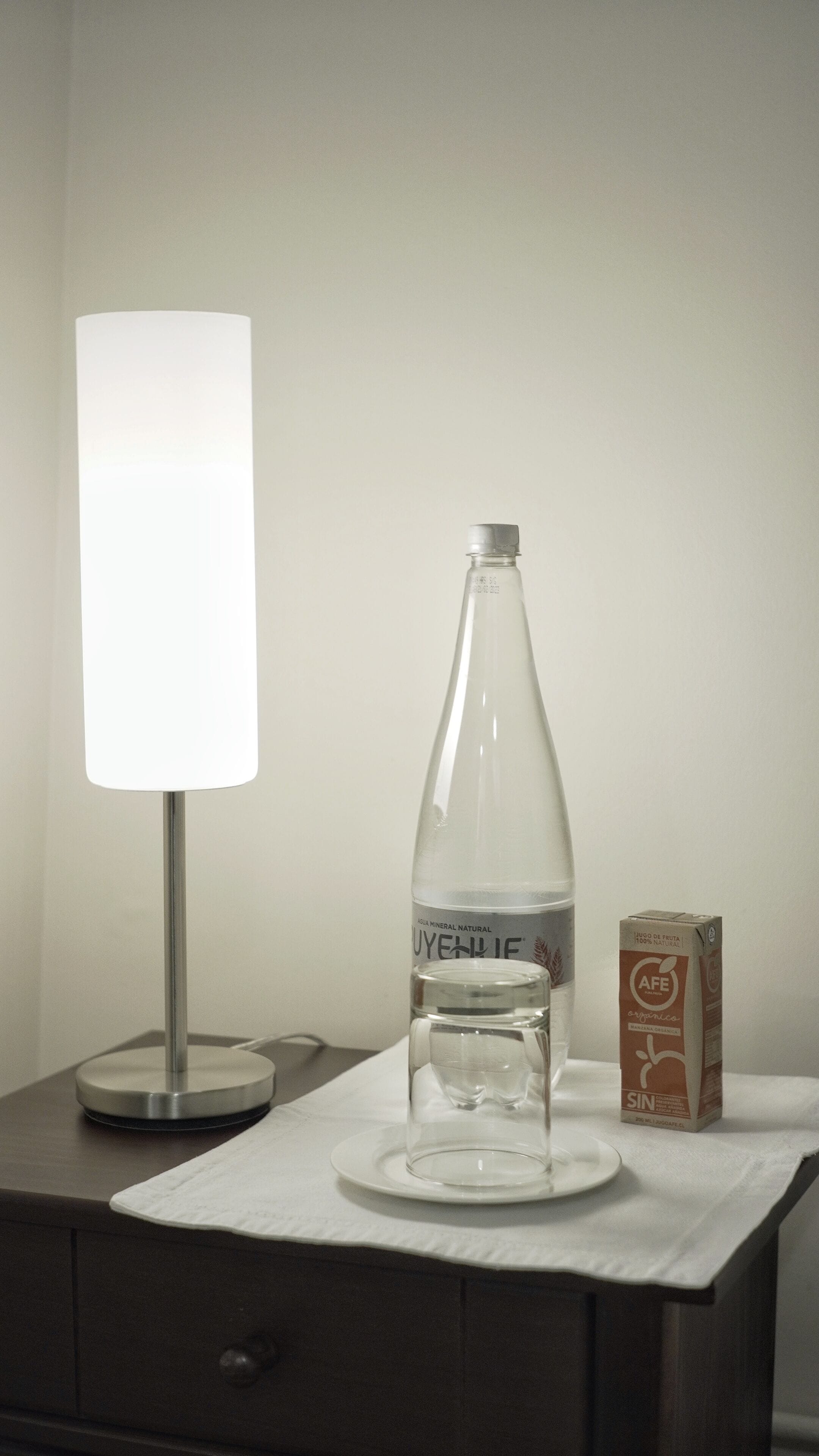 Una mesita de noche con una botella de agua, un vaso y una caja de jugo orgánico sobre un mantel blanco.