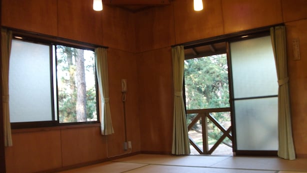 Een kamer met tatami vloerbedekking, houten wanden en grote ramen met gordijnen.