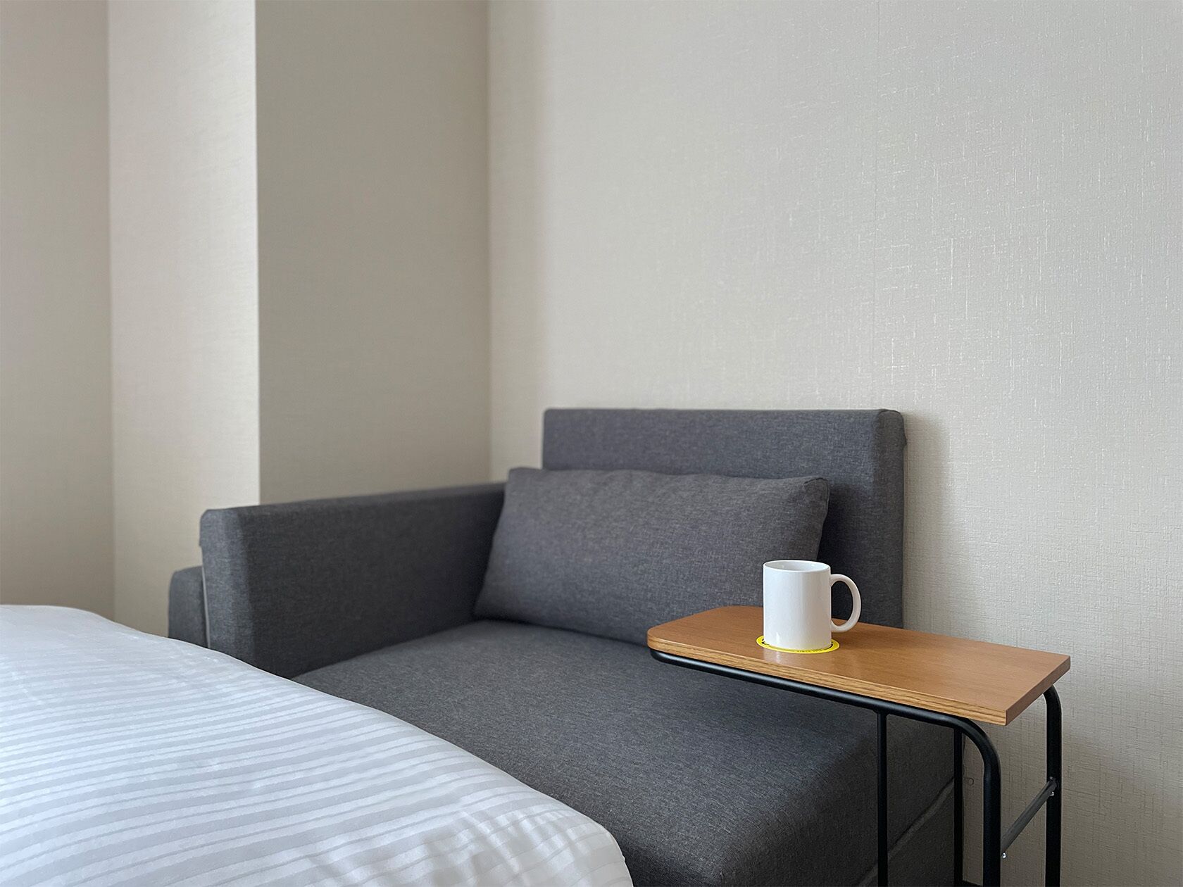 A room with a bed, a grey armchair, and a small wooden table with a white mug.