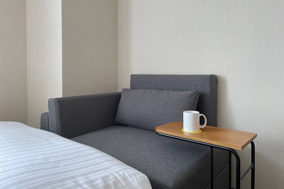 A room with a bed, a grey armchair, and a small wooden table with a white mug.