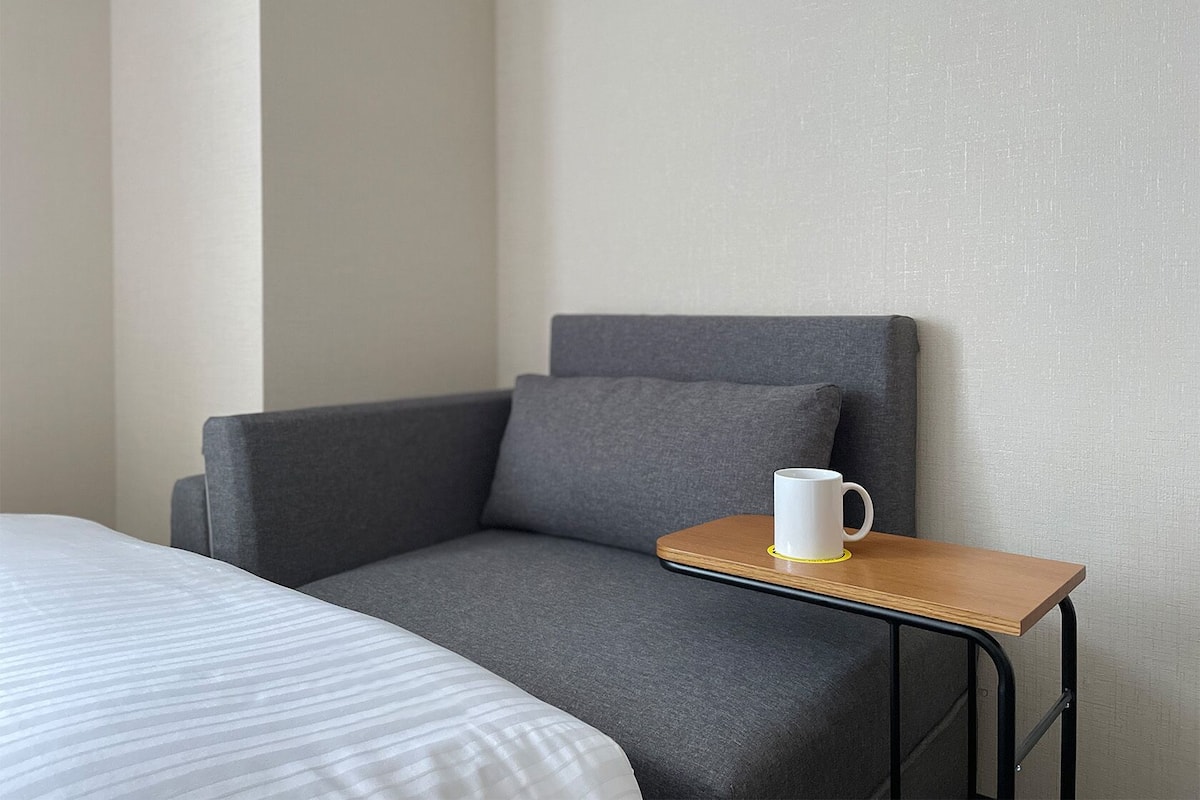 Um quarto com uma cama, uma poltrona cinzenta e uma pequena mesa de madeira com uma caneca branca.