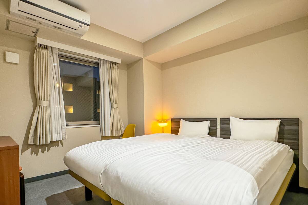 A hotel room with a large bed, a window with curtains, an air conditioning unit, and a bedside lamp.
