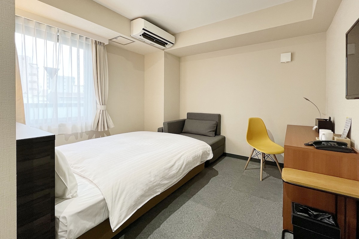 A hotel room with a bed, a desk with a chair, an air conditioning unit, and a window with curtains.