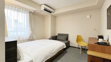 A hotel room with a bed, a desk with a chair, an air conditioning unit, and a window with curtains.