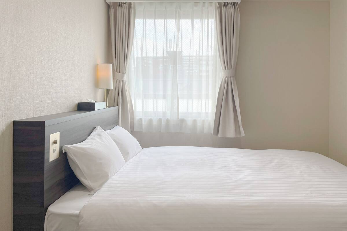 A hotel room with a neatly made bed, a bedside lamp, and a window with sheer curtains.