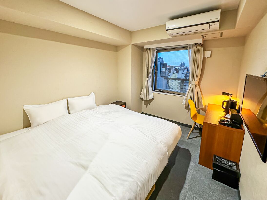 A hotel room with a bed, a desk with a lamp, a chair, and an air conditioning unit.