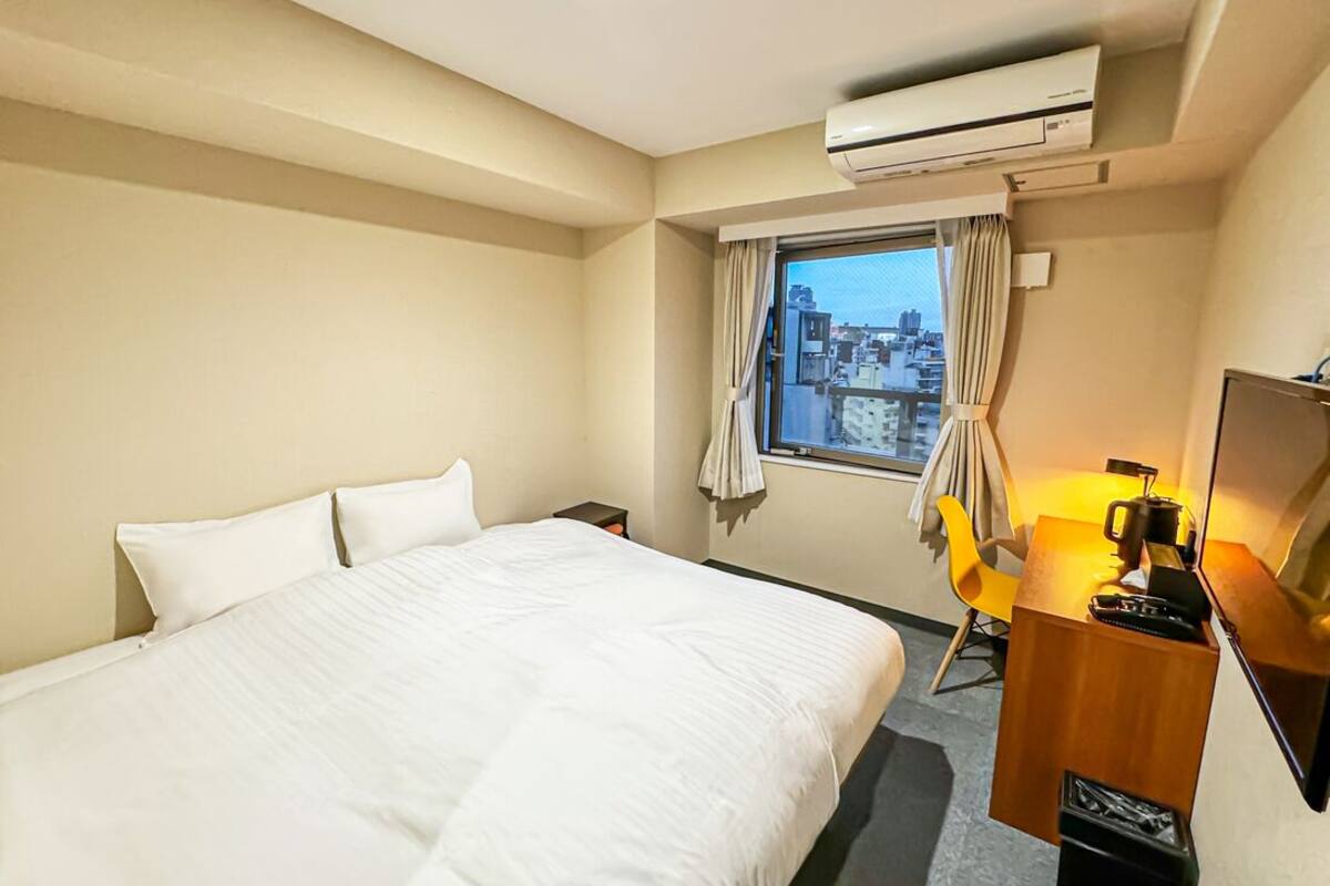 A hotel room with a bed, a desk with a lamp, a chair, and an air conditioning unit.