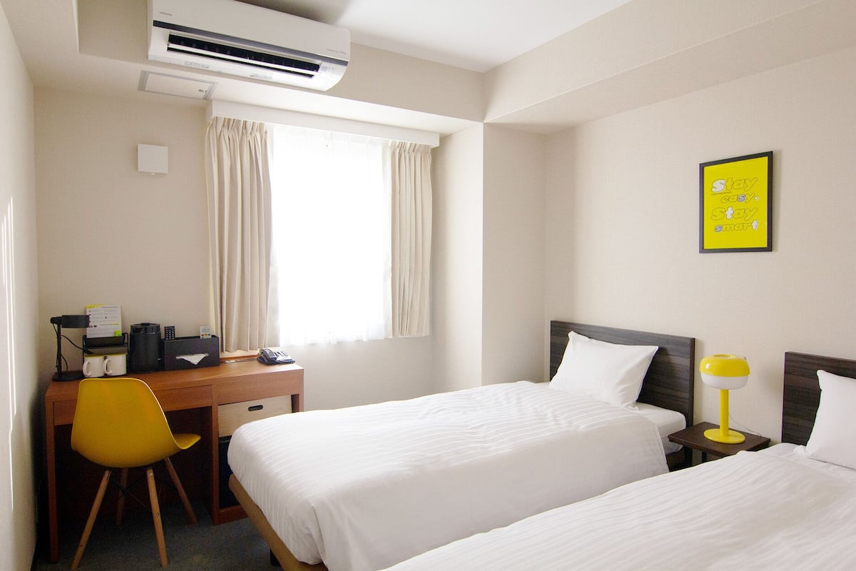 A hotel room with two beds, a desk with a chair, an air conditioner, and a motivational poster on the wall.
