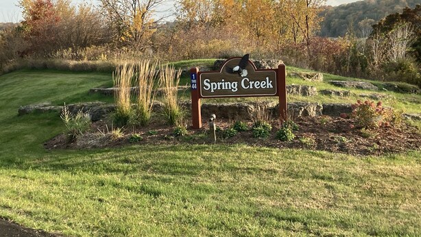 Ein hölzernes Schild mit der Aufschrift 'Spring Creek', umgeben von Gras und Bäumen.