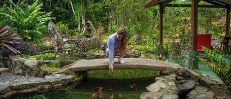 A woman standing on a stone bridge over a pond with koi fish, surrounded by lush greenery and a wooden pavilion.