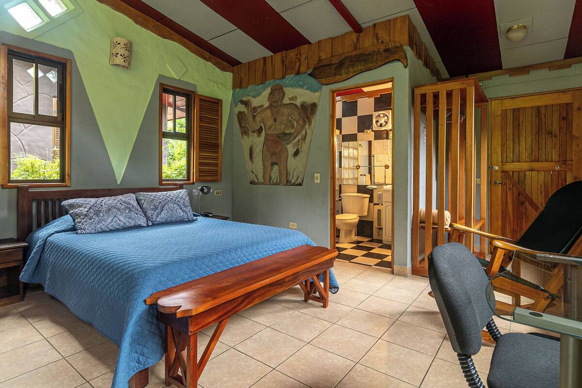 A bedroom with a bed, a bench, a chair, and a view of the bathroom.