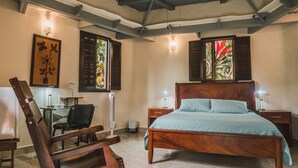 A bedroom with a wooden bed, a desk with a chair, a standing fan, and a window with shutters. - Talamanca Nature Reserve (Rivas)