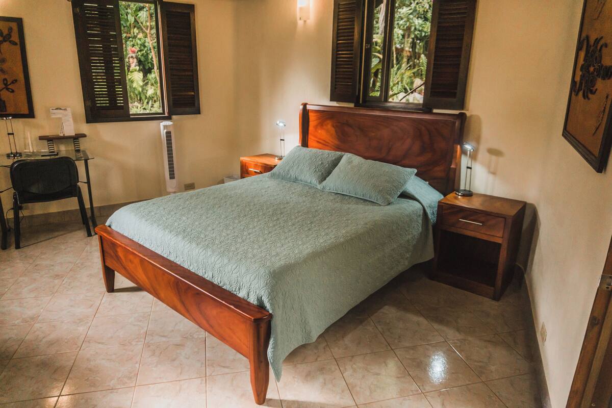 A neatly made bed with a wooden headboard, two bedside tables with lamps, a small desk with a chair, and a window with shutters.