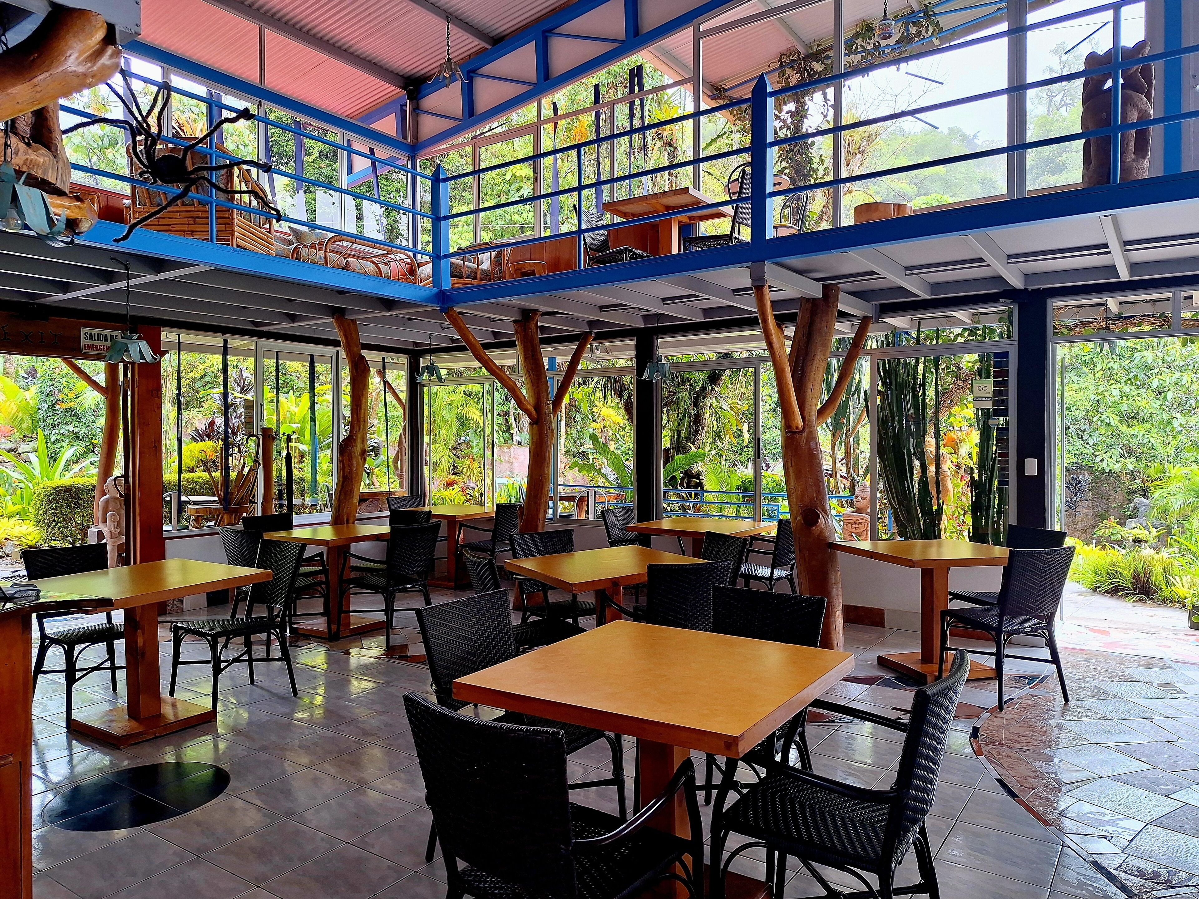 A restaurant with outdoor seating, wooden tables, and chairs, surrounded by large windows and tropical plants.