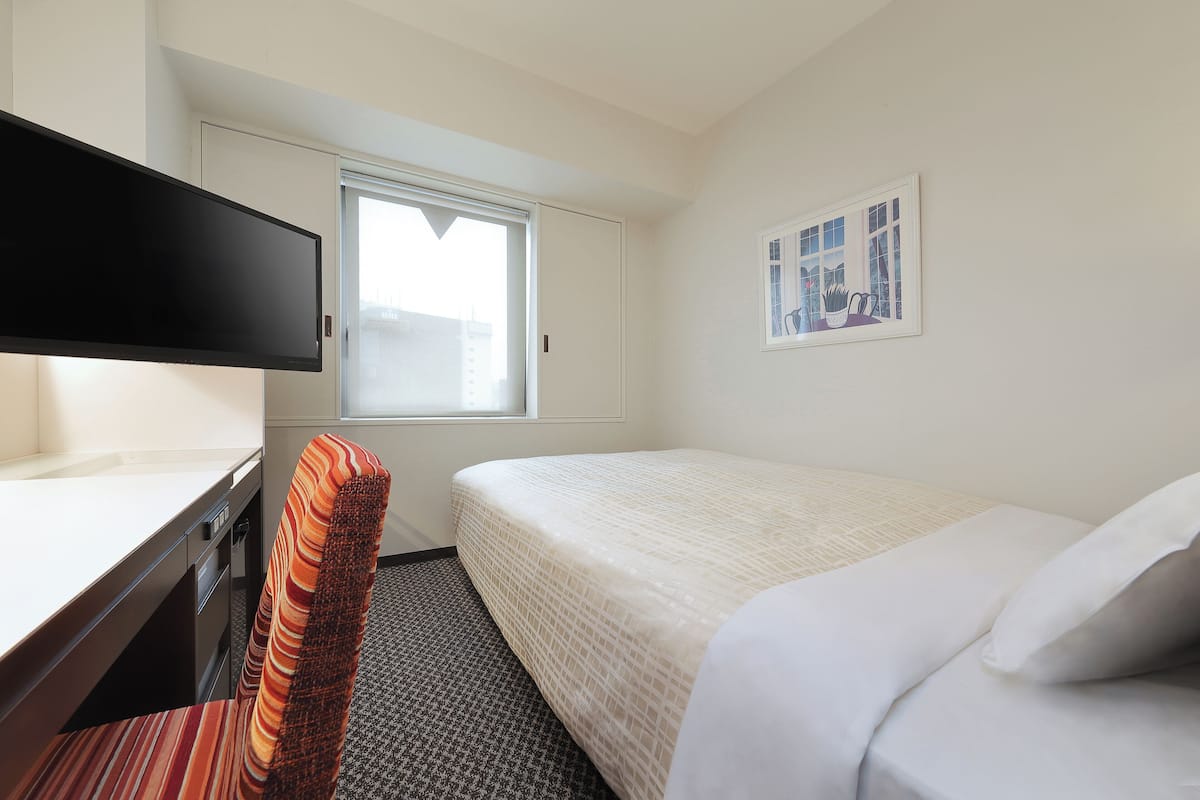 A hotel room with a bed, a desk with a chair, a window, and a framed picture on the wall.