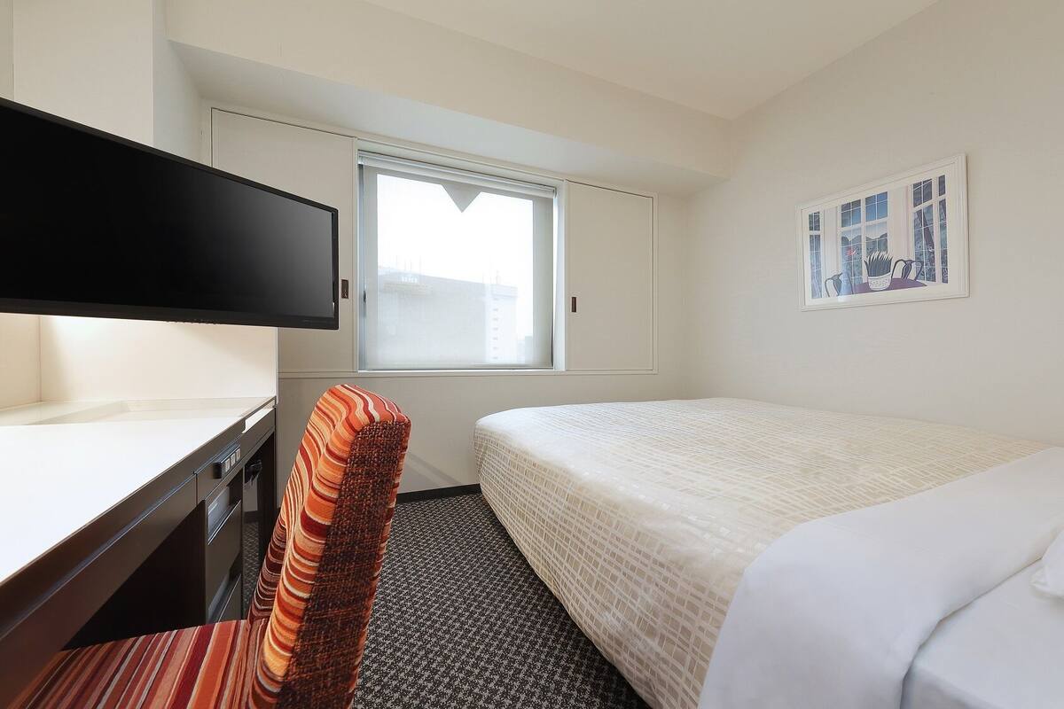 A hotel room with a bed, a desk with a TV, a chair, a window, and a framed picture on the wall.