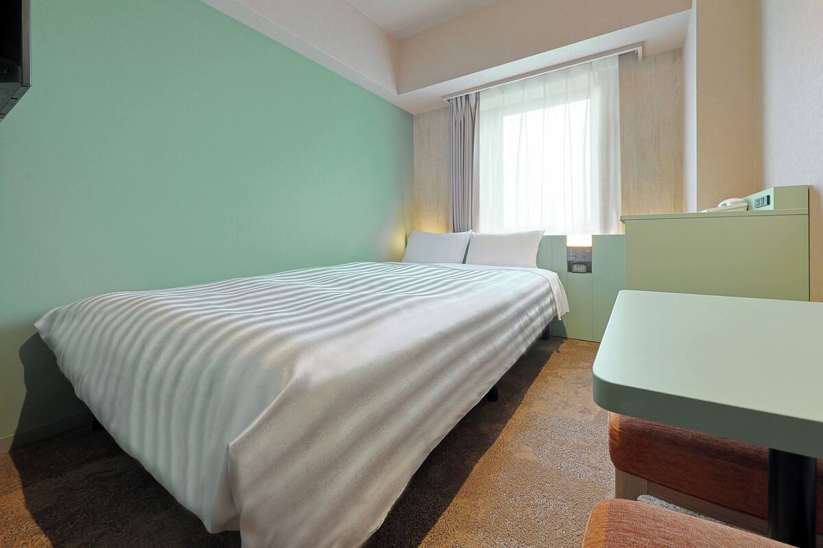 A hotel room with a bed, a desk, a chair, a window with curtains, and a wall clock.