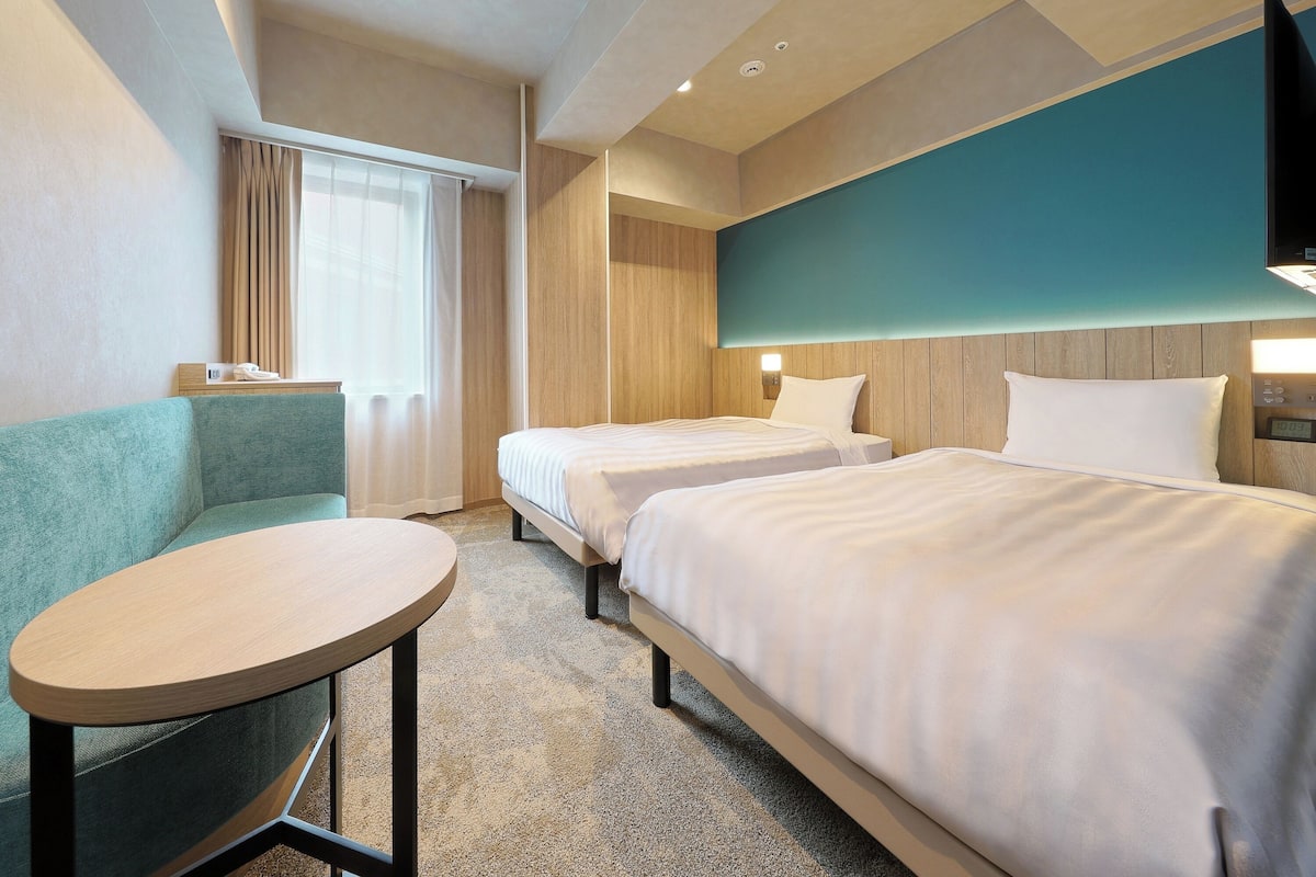A hotel room with two beds, a small round table, a sofa, and a TV mounted on the wall.