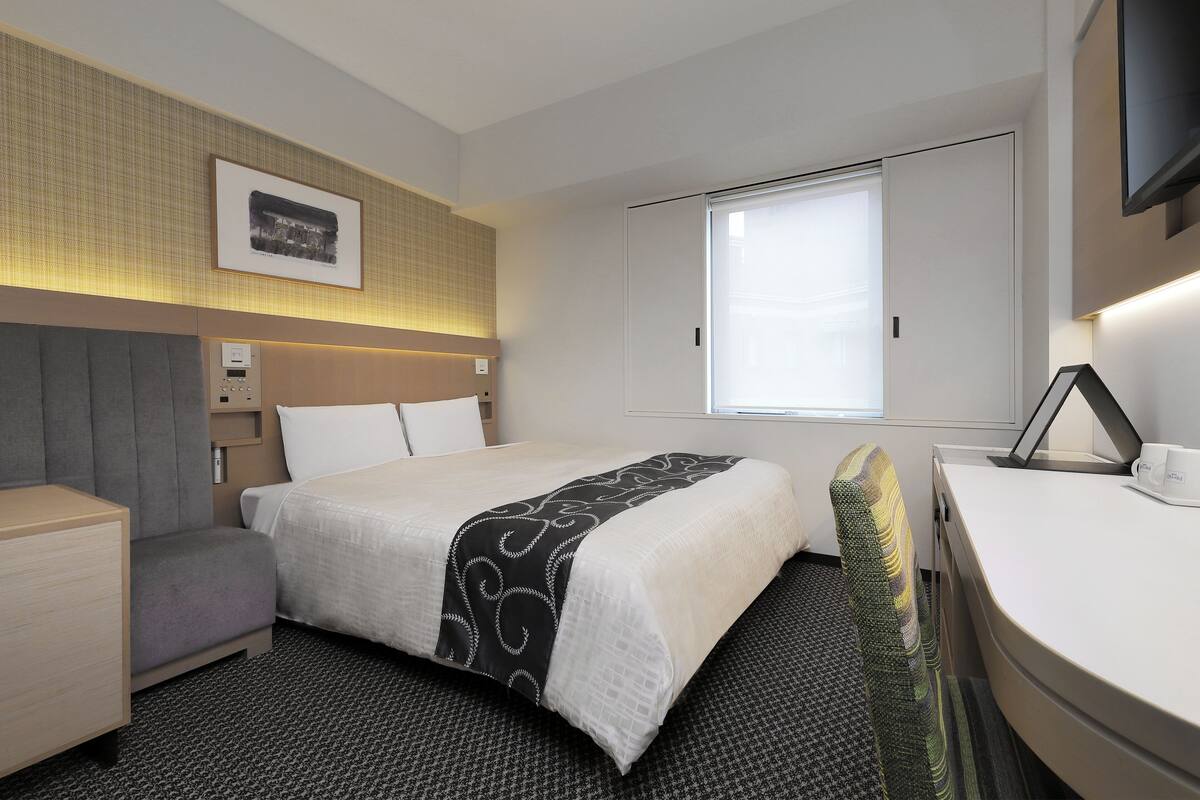 A hotel room with a bed, bedside table, chair, desk, and a television.