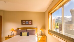A neatly made bed with a wooden headboard, two bedside tables with lamps, a framed picture above the bed, and a large window with a mountain view. - Amazing Rocky Mountain Views: Spacious, Bright, Comfortable Condo, Free WIFI (Canmore)