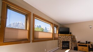 A room with large windows, a stone fireplace, and a television on a wooden stand. - Amazing Rocky Mountain Views: Spacious, Bright, Comfortable Condo, Free WIFI (Canmore)