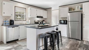 A modern kitchen with white cabinets, a stainless steel refrigerator, a microwave, and a coffee maker. There are three black stools at the island. - On the Canal, by Beach, Kayaks, Dock, Fishing (St. Augustine)