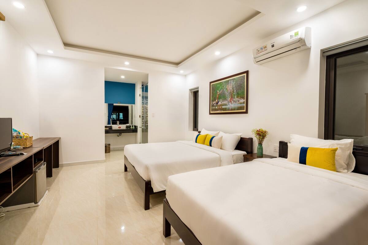 A hotel room with two beds, a desk, a television, and an air conditioning unit.