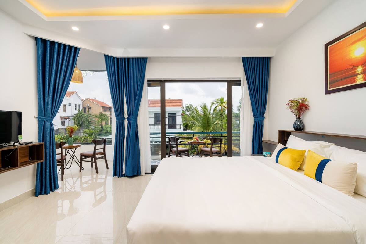A hotel room with a large bed, a TV, a balcony with a table and chairs, and a view of buildings and palm trees.