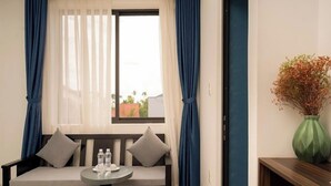 A room with a sofa, a small round table with bottles, a window with sheer curtains, and a vase with dried flowers. - 5 Coconut Homestay Hoi An (Da Nang)