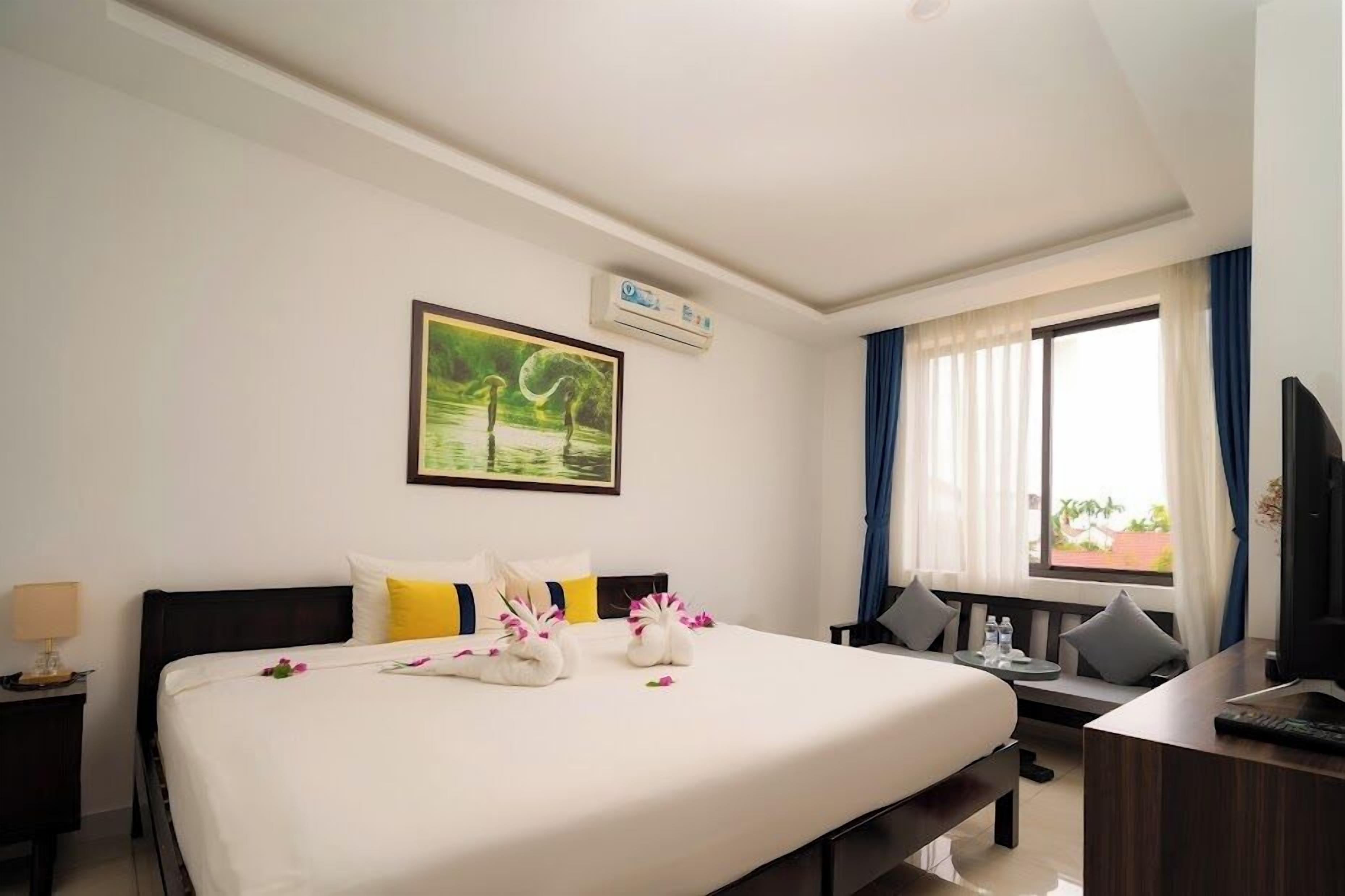 Une chambre d’hôtel avec un grand lit, un bureau avec un ordinateur, une fenêtre avec des rideaux et un tableau au mur.