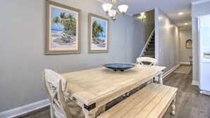 A dining area with a wooden table and bench, two framed beach paintings, a chandelier, and a staircase in the background.