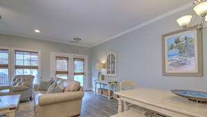 A living room with a beige sofa, a coffee table, and a dining table with benches. There is a lamp on a side table, a framed picture on the wall, and a window with blinds.
