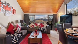 A modern living room with a wooden ceiling, a large window with a mountain view, a sofa with red and beige cushions, a coffee table with a vase of flowers, and a dining area with a table and chairs. - SPACIOUS ALPINE LAKE VIEW SPA TOWNHOUSE WALK TO TOWN (Fernhill)