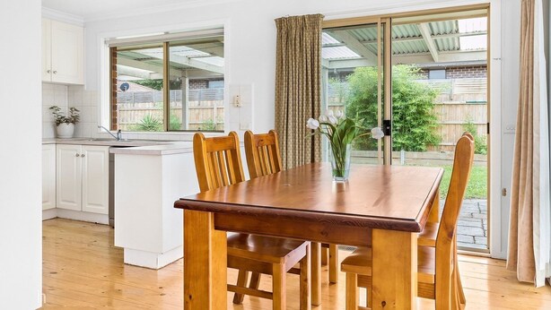 A dining area with a wooden table and chairs, a vase with flowers, and a view of the backyard through large sliding glass doors. - Maroondah 3 Bedroom House (Kilsyth)