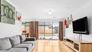 A modern living room with a grey sofa, wooden flooring, a TV mounted on the wall, and a sliding glass door leading to a backyard. - Maroondah 3 Bedroom House (Kilsyth)