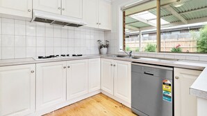 A kitchen with white cabinets, a stainless steel dishwasher, and a window with a view of a backyard. - Maroondah 3 Bedroom House (Kilsyth)