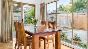 A dining area with a wooden table and chairs, large windows, and a view of a backyard with a wooden fence and greenery. - Maroondah 3 Bedroom House (Kilsyth)