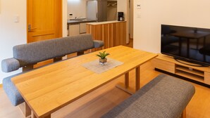 Una sala de estar moderna con una mesa de comedor de madera, dos sofás grises, un banco, un televisor de pantalla plana y una cocina al fondo.