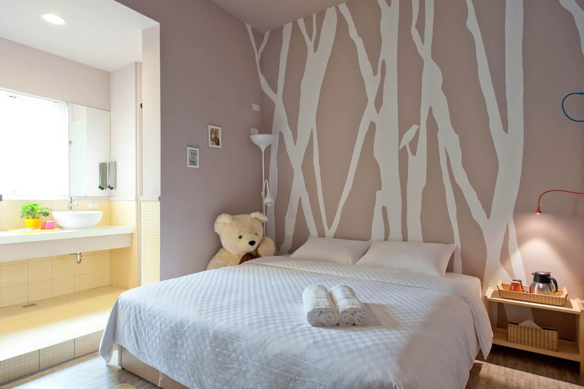 Une chambre à coucher avec un lit, une table de chevet avec une lampe, un ours en peluche, et une salle de bain avec un lavabo et un miroir.